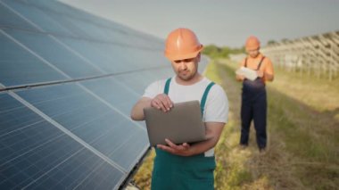 Güneş pili panelleriyle ayakta duran mühendis güneş hücresinden üretilen elektrik enerjisinin değerinin monitörüne bakıyor. Dizüstü bilgisayarı elinde tutan adam güneş panelleriyle sahada duruyor..