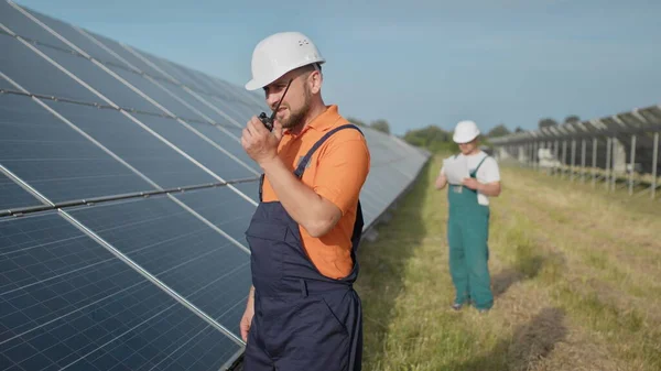 Um empregado de uma central eléctrica transmite comandos por walkie-talkie. Engenheiro adulto caucasiano feliz no capacete sorrindo para a câmera na estação de energia solar fora. Indústria ecológica. Equipa empresarial — Fotografia de Stock