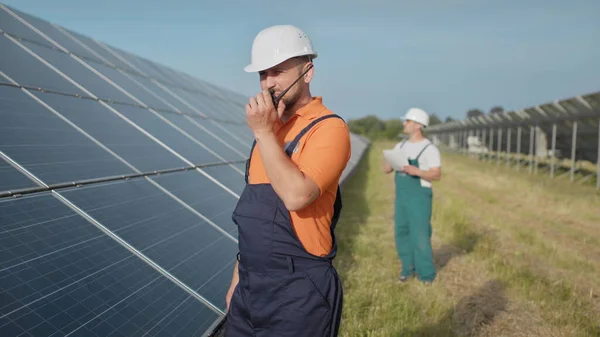 En ansatt ved et kraftverk sender kommandoer via walkie-talkie. Happy caucasian voksen ingeniør i hjelm smilende foran kamera på solstasjonen utenfor. Økologisk industri. Forretningslag – stockfoto