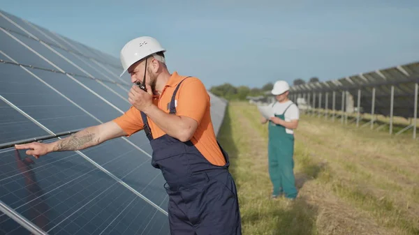 Um empregado de uma central eléctrica transmite comandos por walkie-talkie. Engenheiro adulto caucasiano feliz no capacete sorrindo para a câmera na estação de energia solar fora. Indústria ecológica. Equipa empresarial — Fotografia de Stock