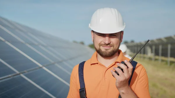 Engenheiro adulto caucasiano feliz no capacete sorrindo para a câmera na estação de energia solar fora. Indústria ecológica. Conceito de fazenda solar. Um funcionário de uma usina transmite comandos por walkie-talkie — Fotografia de Stock