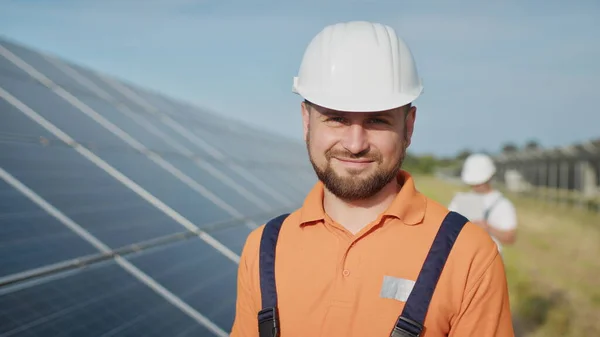Engenheiro adulto caucasiano feliz no capacete sorrindo para a câmera na estação de energia solar fora. Indústria ecológica. Conceito de fazenda solar. Um funcionário de uma usina transmite comandos por walkie-talkie — Fotografia de Stock