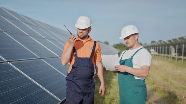 Aurinkovoimalan insinöörit. Aikuiset miehet kypärissä, Voimalaitoksen työntekijä lähettää käskyjä radiopuhelimella. Puhtaan energian kuljetus. Puhdasta energiaa. Aurinkoparistot. — kuvapankkivalokuva