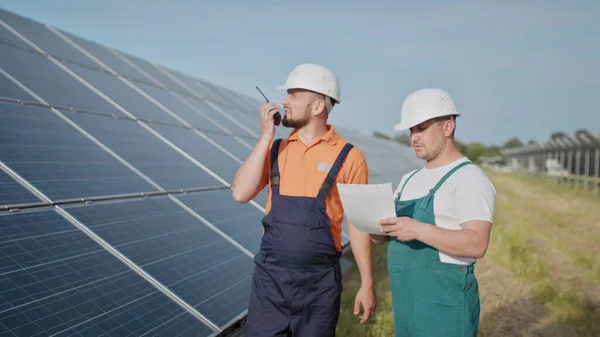 Aurinkovoimalan insinöörit. Aikuiset miehet kypärissä, Voimalaitoksen työntekijä lähettää käskyjä radiopuhelimella. Puhtaan energian kuljetus. Puhdasta energiaa. Aurinkoparistot. — kuvapankkivalokuva