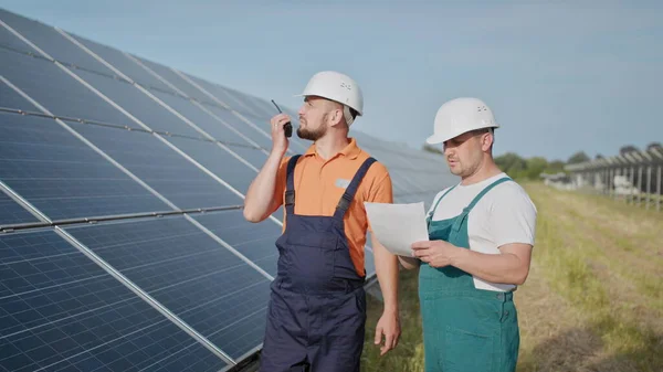 Aurinkovoimalan insinöörit. Aikuiset miehet kypärissä, Voimalaitoksen työntekijä lähettää käskyjä radiopuhelimella. Puhtaan energian kuljetus. Puhdasta energiaa. Aurinkoparistot. — kuvapankkivalokuva