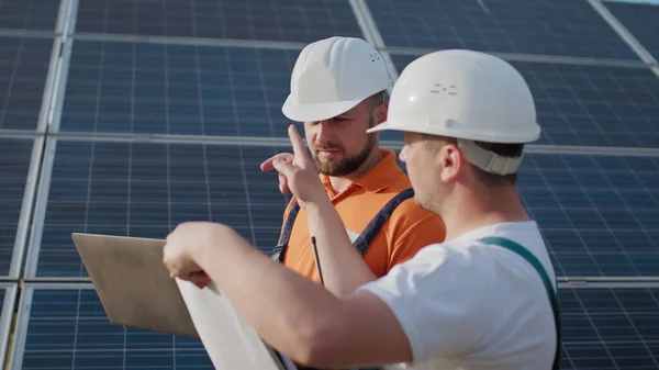 Lähikuva insinöörit käyttävät kannettavaa tietokonetta ja projektisuunnitelma aurinkopaneeli erityinen yhtenäinen seisoo katsoa ympärilleen keskustella asennus aurinkoinen akkujen sähkön ympäristöön. Vihreän energian työpaikat — kuvapankkivalokuva
