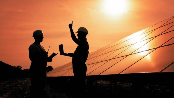 Two Unidentified Solar Power Engineers In Backlight At Sunset. Team of industrial colleagues using tablet monitoring rows of photovoltaic solar panels at sunset. Solar park. Alternative energy concept