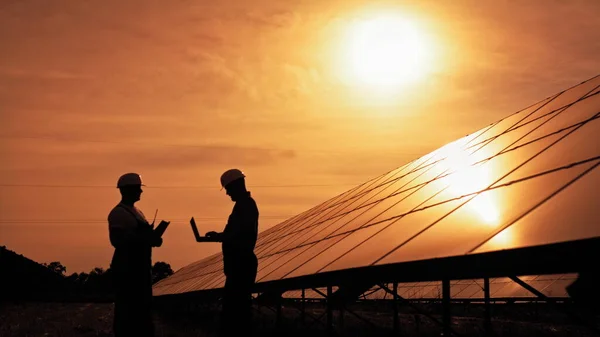 Dwóch niezidentyfikowanych inżynierów energii słonecznej rozmawia ze sobą o zachodzie słońca. Kilku pracowników badających baterie słoneczne i komunikujących się w interesach. Ekologiczna przyszłość. Zrównoważona energia. — Zdjęcie stockowe