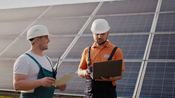 Tekniker med bærbar PC som sjekker panelene ved solenergiinstallasjonen. Forretningssamarbeid. Solpark. Alternativt energibegrep. Ren energi. To arbeidere i uniform og hatt – stockfoto