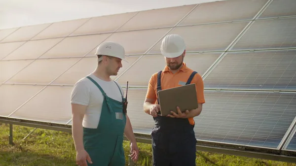 วิศวกรชายยืนที่ฟาร์มพลังงานแสงอาทิตย์และหารือเกี่ยวกับแผนการก่อสร้างที่มีประสิทธิภาพ ชายสองคนสวมหมวกป้องกัน วิศวกรจับมือกันในอากาศ สถานที่สําหรับเครื่องบันทึกหน้าจอเสมือน . — ภาพถ่ายสต็อก