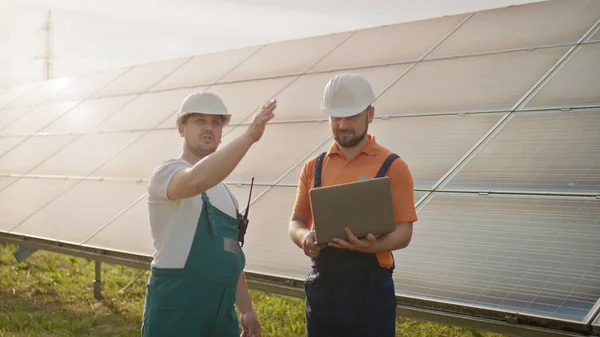 Mannlige ingeniører som står på solfarmen og diskuterer effektive byggeplaner. To menn i beskyttelseshjelm. Ingeniøren holder hånden i været. Et sted for en virtuell skjermsparer.. – stockfoto