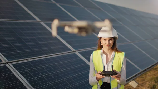 Engineer woman in hardhat holding tablet computer operating flying drone in solar plant. Photovoltaic solar panel installation. Solar array. New technologies. The investor checks the solar power