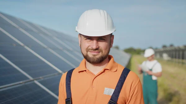 Šťastný kavkazský dospělý inženýr v helmě, usmívající se na kameru před solární elektrárnou. Ekologický průmysl. Koncept solární farmy. Zaměstnanec elektrárny přenáší příkazy pomocí vysílačky. Stock Obrázky