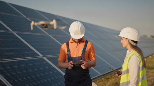 Teollisuuden asiantuntija yllään kypärä ja ohjaus drone aurinkosähkövoimalassa. Aurinkopaneelien asennus. Teknologiat ja ekologia. Naissijoittaja tarkastaa työn — kuvapankkivalokuva