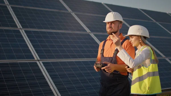 Industrial expert wearing helmet and controlling drone in photovoltaic solar power plant. Solar panel array installation. Technologies and ecology. Female investor checks the work