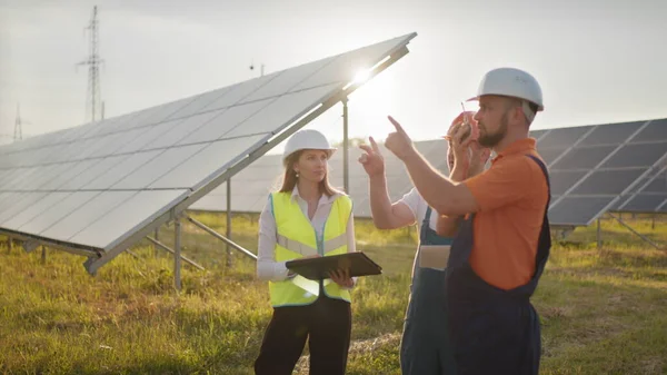 Kolme aurinkoenergian asiantuntijaa aurinkovoimalassa. Ammattitaitoiset insinöörit keskustelevat innovatiivisesta hankkeesta. Teollisuusteknikoiden tiimi aurinkopuistossa. Aurinkovoimalan rakentaminen. — kuvapankkivalokuva