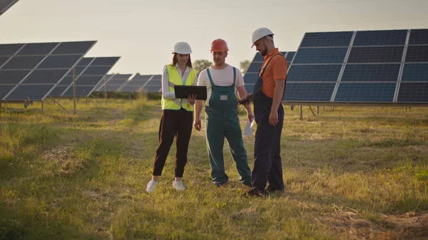 ผู้เชี่ยวชาญด้านพลังงานแสงอาทิตย์สามคน ที่โรงไฟฟ้าพลังงานแสงอาทิตย์ เด็กเล็กสองคนวิ่งไปหาพ่อแม่ที่ทํางาน พนักงานโรงไฟฟ้าพลังงานแสงอาทิตย์ที่มีเด็กในที่ทํางานยิ้มที่กล้อง แฮปปี้ — ภาพถ่ายสต็อก