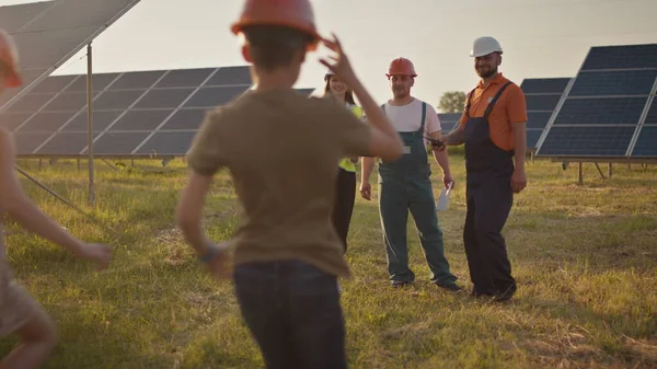 Three solar energy specialists at a solar power facility. Two small children run to their parents who are at work. A solar power plant employee with children at work smiling at the camera. Happy