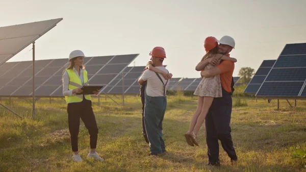 Tre spesialister på solenergi ved et solkraftverk. To små barn løper til foreldrene som er på jobb. En ansatt i et solkraftverk med barn på jobb som smiler til kameraet. Lykkelig – stockfoto