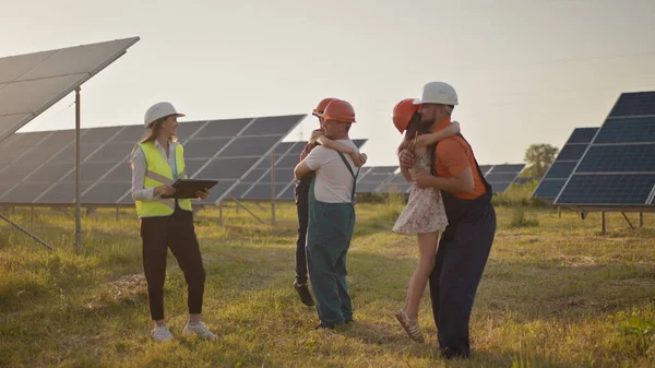 Tre spesialister på solenergi ved et solkraftverk. To små barn løper til foreldrene som er på jobb. En ansatt i et solkraftverk med barn på jobb som smiler til kameraet. Lykkelig – stockfoto