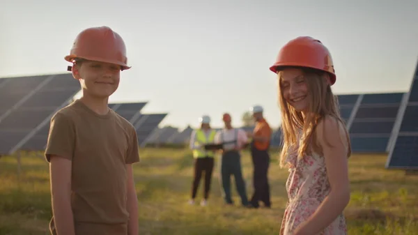 Duas crianças pequenas correm para os pais que estão no trabalho. Um funcionário da usina solar com crianças no trabalho sorrindo para a câmera. Três especialistas em energia solar em uma usina de energia solar. — Fotografia de Stock