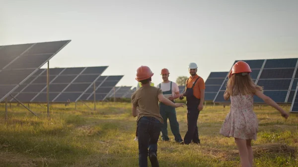 เด็กเล็กสองคนวิ่งไปหาพ่อแม่ที่ทํางาน พนักงานโรงไฟฟ้าพลังงานแสงอาทิตย์ที่มีเด็กในที่ทํางานยิ้มที่กล้อง ผู้เชี่ยวชาญด้านพลังงานแสงอาทิตย์สามคนที่โรงไฟฟ้าพลังงานแสงอาทิตย์ . — ภาพถ่ายสต็อก