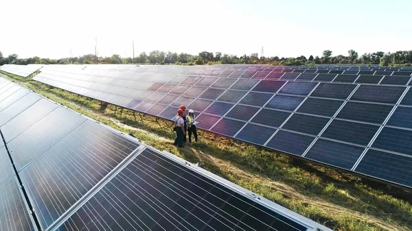 Kolme aurinkoenergian asiantuntijaa kävelee aurinkovoimalassa. Ammattitaitoiset insinöörit keskustelevat innovatiivisesta hankkeesta. Ilmakuvamateriaalia satoja aurinkoenergiamoduuleja tai paneeleja. Työtovereiden ryhmä — kuvapankkivalokuva