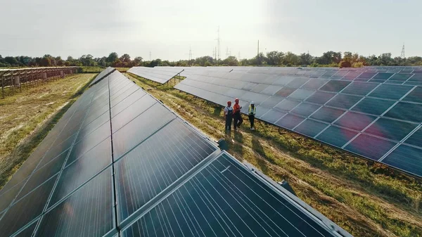 Kolme aurinkoenergian asiantuntijaa kävelee aurinkovoimalassa. Ammattitaitoiset insinöörit keskustelevat innovatiivisesta hankkeesta. Top näkymä asiantuntijoiden kävely yli aurinkovoimalan. — kuvapankkivalokuva