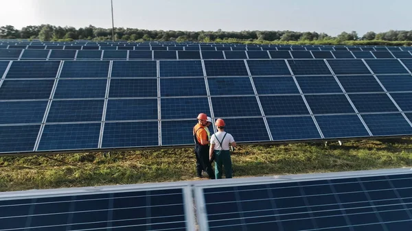 Ilmakuva kolmesta aurinkoenergiainsinööristä suurella aurinkotilalla. Kolme vaihtoehtoisen voimalan työntekijää kävelee ja puhuu aurinkopaneelien järjestelmästä.Teknikko ja sijoittaja — kuvapankkivalokuva