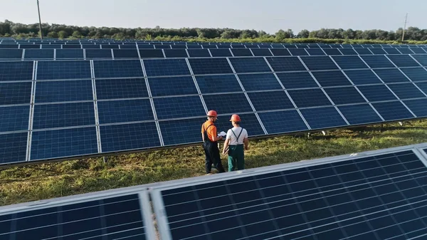 Tiro aéreo de três engenheiros de energia solar em uma grande fazenda solar. Três funcionários de usina de energia alternativa andando e falando sobre esquema de painéis solares. Técnico e investidor — Fotografia de Stock