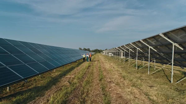 Zdjęcie lotnicze trzech inżynierów energii słonecznej na dużej farmie słonecznej. Trzech pracowników elektrowni alternatywnych spacerujących i mówiących o schemacie paneli słonecznych. Ludzie są zadowoleni z sukcesu pracy — Zdjęcie stockowe