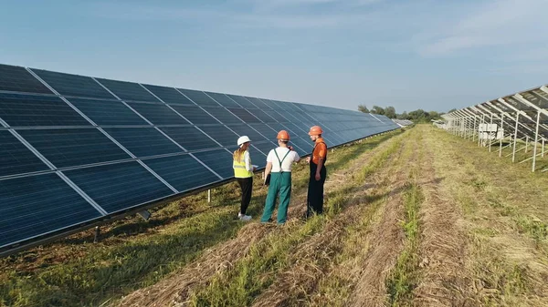 Tre ansatte ved alternativt kraftverk som går og snakker om solcelleordning. Vedlikeholdslag med en bærbar datamaskin observerer et solkraftfelt. Solkraft, solcellepaneler, solpark – stockfoto