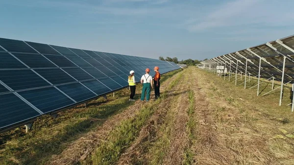 Trois employés de la centrale alternative marche et parler de schéma de panneaux solaires. Équipe commerciale d'ingénieurs industriels marchant sur la ferme solaire et discutant du plan efficace de la construction. — Photo