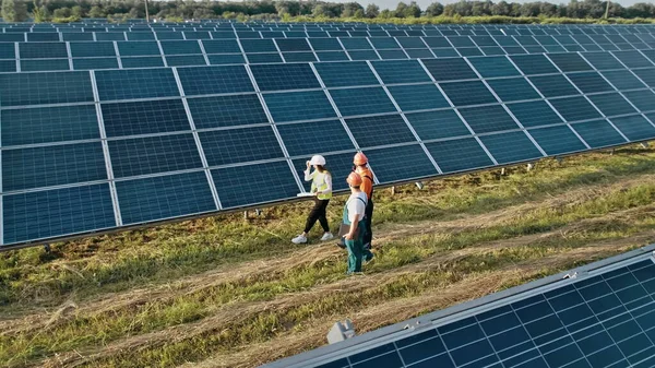 Kolme vaihtoehtoisen voimalaitoksen työntekijää kävelee ja puhuu aurinkopaneelien järjestelmästä. Teknikko ja sijoittaja kävely Solar cell Farm läpi kentän aurinkopaneelit tarkkailun Solar — kuvapankkivalokuva