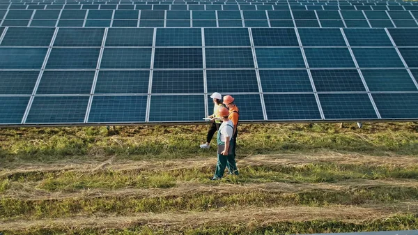 Kolme vaihtoehtoisen voimalaitoksen työntekijää kävelee ja puhuu aurinkopaneelien järjestelmästä. Teknikko ja sijoittaja kävely Solar cell Farm läpi kentän aurinkopaneelit tarkkailun Solar — kuvapankkivalokuva