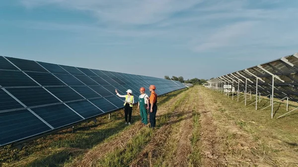 Equipe de negócios de engenheiros industriais caminhando na fazenda solar e discutindo plano eficiente de construção. Três funcionários de usina de energia alternativa andando e falando sobre esquema de painéis solares. — Fotografia de Stock