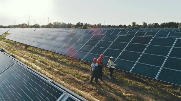 Top näkymä asiantuntijoiden kävely yli aurinkovoimalan. Kolme vaihtoehtoisen voimalan työntekijää kävelee ja puhuu aurinkopaneelien järjestelmästä. — kuvapankkivalokuva