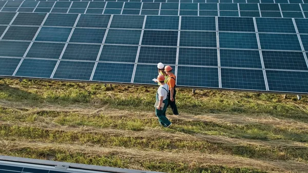 Kolme vaihtoehtoisen voimalaitoksen työntekijää kävelee ja puhuu aurinkopaneelien järjestelmästä. Teknikko ja sijoittaja kävely Solar cell Farm läpi kentän aurinkopaneelit tarkkailun Solar tekijänoikeusvapaita valokuvia kuvapankista