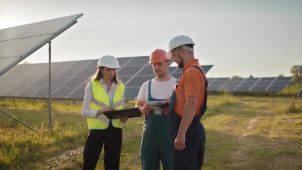 Trois spécialistes de l'énergie solaire dans une centrale solaire. Des ingénieurs professionnels discutent de projets innovants. Équipe de techniciens industriels dans le parc solaire. Construction d'une centrale solaire. — Video