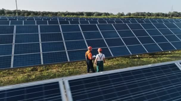 Повітряний постріл трьох інженерів сонячної енергії на великій сонячній фермі. Три співробітники альтернативної електростанції ходять і говорять про схему сонячних панелей. Технік та інвестор — стокове відео