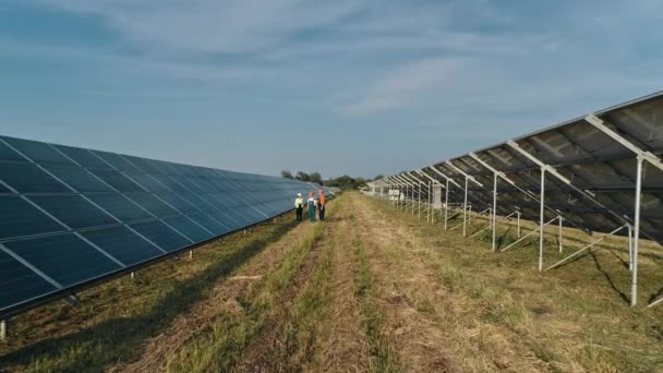 Εναέρια λήψη τριών μηχανικών ηλιακής ενέργειας σε ένα μεγάλο ηλιακό αγρόκτημα. Τρεις υπάλληλοι της εναλλακτικής μονάδας παραγωγής ενέργειας περπατώντας και μιλώντας για το σχήμα των ηλιακών συλλεκτών. Οι άνθρωποι είναι ικανοποιημένοι επιτυχία στην εργασία — Αρχείο Βίντεο