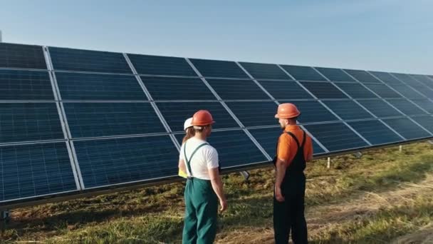 Três funcionários de usina de energia alternativa andando e falando sobre o esquema de painéis solares. Manutenção O trabalho em equipe com um laptop está observando um campo de energia solar. Energia solar, painel solar, fazenda solar — Vídeo de Stock