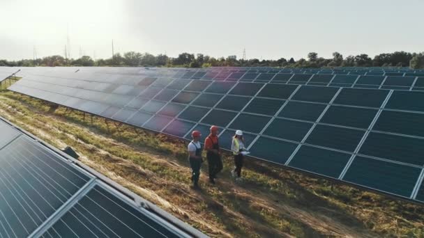 Blick von oben auf Spezialisten, die über ein Solarkraftwerk gehen. Drei Mitarbeiter des alternativen Kraftwerks gehen und sprechen über das System der Sonnenkollektoren. — Stockvideo