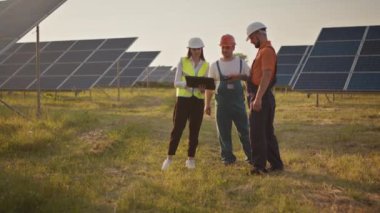 Bir güneş enerjisi tesisinde üç güneş enerjisi uzmanı. İki küçük çocuk iş yerindeki ebeveynlerine koşuyor. İş yerinde çocukları kameraya gülümseyen bir güneş enerjisi santrali çalışanı. Mutlu