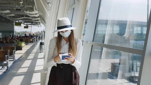 Mulher de negócios bonita no aeroporto, usando o telefone para enviar mensagens de texto, em pé junto à janela no prédio do escritório corporativo, olhando para a câmera. A menina está segurando bilhetes e um — Fotografia de Stock