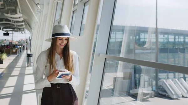 Elegant beautiful business girl holding travel tickets and using phone, happy girl looking at camera in airport terminal. Fashion woman portrait. Airport terminal. Business trip