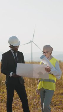 İki mühendis, biri başlık takıyor, diğeri de tesettür. Sürdürülebilir enerjiyi temsil eden rüzgar türbinlerinin önündeki planları inceliyorlar..