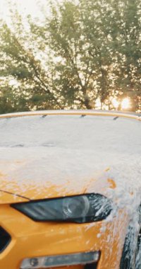 Sarı bir arabanın köpüklü sabunla yıkandığı dikey bir çekim, elinde bir basınç makinası tutan bir kişi..