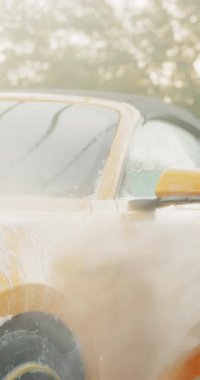 Sarı bir arabanın dikey görüntüsü yıkanırken, ön cam ve gövdeye su püskürtülüyor..