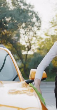 Sarı bir arabanın ön camını yeşil bir bezle temizleyen bir kişi görülüyor. Ağaçların ve güneş ışığının bulanık arkaplanı var..